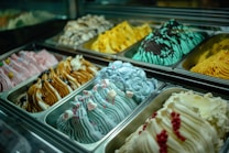 Rows of colorful gelato flavors are displayed in metal trays. Each flavor has a unique texture and color, with decorative toppings like berries, marshmallows, and sprinkles. The lighting highlights the vibrant colors and creamy texture of the gelato.