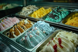 Rows of colorful gelato flavors are displayed in metal trays. Each flavor has a unique texture and color, with decorative toppings like berries, marshmallows, and sprinkles. The lighting highlights the vibrant colors and creamy texture of the gelato.