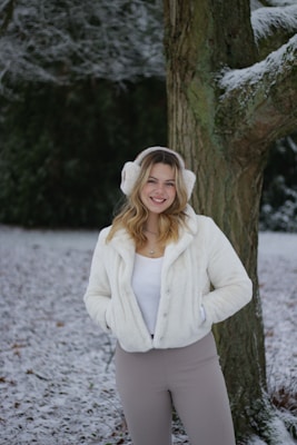A woman is standing in a snowy landscape, wearing a white fuzzy jacket and earmuffs. She has blonde hair and is smiling, with her hands in the pockets of her jacket. Behind her, there is a large tree trunk dusted with snow and a dark green background suggesting forest or shrubs.