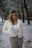 A woman with light brown hair stands in a snowy park, wearing a white fur jacket, white top, and beige pants. She holds a fur-lined accessory in her left hand while facing the camera directly. The background features leafless trees and a snow-covered path, creating a winter scene.