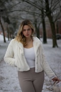 A woman with light brown hair stands in a snowy park, wearing a white fur jacket, white top, and beige pants. She holds a fur-lined accessory in her left hand while facing the camera directly. The background features leafless trees and a snow-covered path, creating a winter scene.