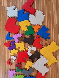A collection of interlocking plastic bricks assembled into various abstract shapes lies scattered on a wooden floor. The shapes are made up of different colors and sizes, creating a vibrant, patchwork-like appearance.