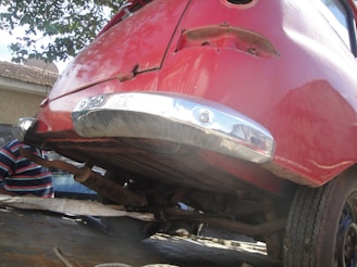 The image depicts the rear section of an old car elevated on a platform or incline. The vehicle is painted in a faded red color with some visible rust and deterioration. The chrome bumper appears reflective but has signs of wear. Part of a human figure in a striped shirt is partially visible in the background amid an outdoor setting.