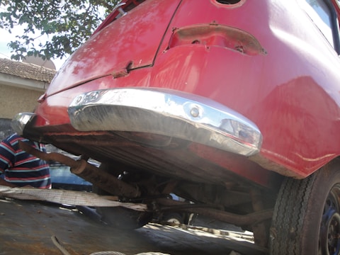 The image depicts the rear section of an old car elevated on a platform or incline. The vehicle is painted in a faded red color with some visible rust and deterioration. The chrome bumper appears reflective but has signs of wear. Part of a human figure in a striped shirt is partially visible in the background amid an outdoor setting.