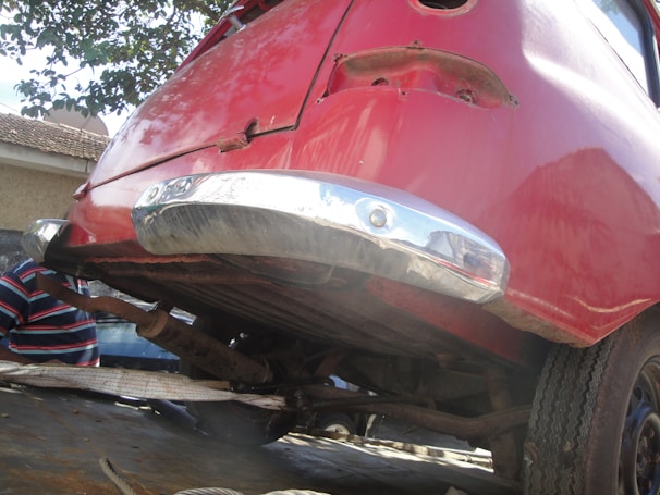 The image depicts the rear section of an old car elevated on a platform or incline. The vehicle is painted in a faded red color with some visible rust and deterioration. The chrome bumper appears reflective but has signs of wear. Part of a human figure in a striped shirt is partially visible in the background amid an outdoor setting.