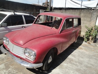 An old, dusty red car is parked in a concrete area beside a white vehicle. The car appears to be vintage, with a noticeable lack of modern features. The surroundings include a brick wall and some vegetation growing nearby.