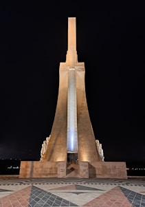 A tall monument with a central vertical structure illuminated at night, standing against a dark sky. It features symmetrical design elements flanked by sculptures on either side. The base includes steps leading up to the monument, with patterned tiles extending in front.