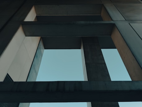 A perspective view of a concrete architectural structure with intersecting beams and pillars. The design features sharp angles and clean lines, framed by the open sky visible through the gaps between the structures.