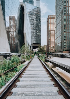 A perspective view of an urban scene where old railway tracks are flanked by green plants, leading the eye towards modern skyscrapers with reflective glass facades. The area appears to be a reused industrial space, now transformed into a park-like passageway amidst the towering city buildings.