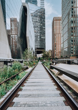 A perspective view of an urban scene where old railway tracks are flanked by green plants, leading the eye towards modern skyscrapers with reflective glass facades. The area appears to be a reused industrial space, now transformed into a park-like passageway amidst the towering city buildings.