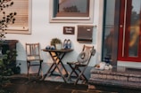 A cozy outdoor seating area featuring two wooden chairs and a small round table. On the table, there is a decorative plant and a pair of blue clogs with a floral pattern. A rustic wreath with a heart hangs from one chair. A stone patio and a red door are adjacent, with a mailbox and a sign hanging on the wall. Various items like a thermos, a candle, and cups are placed around.