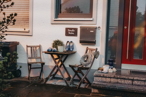 A cozy outdoor seating area featuring two wooden chairs and a small round table. On the table, there is a decorative plant and a pair of blue clogs with a floral pattern. A rustic wreath with a heart hangs from one chair. A stone patio and a red door are adjacent, with a mailbox and a sign hanging on the wall. Various items like a thermos, a candle, and cups are placed around.
