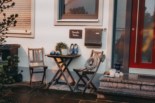 A cozy outdoor seating area featuring two wooden chairs and a small round table. On the table, there is a decorative plant and a pair of blue clogs with a floral pattern. A rustic wreath with a heart hangs from one chair. A stone patio and a red door are adjacent, with a mailbox and a sign hanging on the wall. Various items like a thermos, a candle, and cups are placed around.