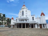 A large white church with red roof tiles and a central cross at the top. It features a symmetrical façade with multiple decorative elements, including stained glass windows and arched doorways. Palms and other buildings are visible in the background.