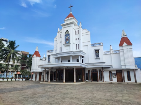 A large white church with red roof tiles and a central cross at the top. It features a symmetrical façade with multiple decorative elements, including stained glass windows and arched doorways. Palms and other buildings are visible in the background.
