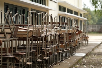 A row of wooden school desks and chairs stacked upside down outside a building with large windows. The setting appears to be outdoors, and the area around the building is clear and orderly.