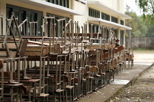 A row of wooden school desks and chairs stacked upside down outside a building with large windows. The setting appears to be outdoors, and the area around the building is clear and orderly.