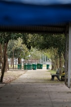 A series of green trash bins lined up along a concrete path, surrounded by trees and foliage. There is a wooden bench on the right side, and the path is partially shaded by a roof structure. The scene is set in a park-like area with natural elements such as trees and grass.