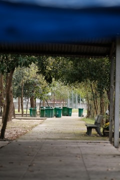 A series of green trash bins lined up along a concrete path, surrounded by trees and foliage. There is a wooden bench on the right side, and the path is partially shaded by a roof structure. The scene is set in a park-like area with natural elements such as trees and grass.