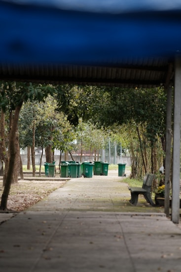 A series of green trash bins lined up along a concrete path, surrounded by trees and foliage. There is a wooden bench on the right side, and the path is partially shaded by a roof structure. The scene is set in a park-like area with natural elements such as trees and grass.