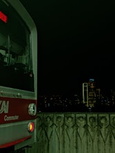 A commuter train is visible on the left side against a dark nighttime cityscape. The train has a red and silver exterior with a digital display showing text in red. The foreground features a patterned concrete barrier, and in the background, city buildings are faintly illuminated with a few bright lights.