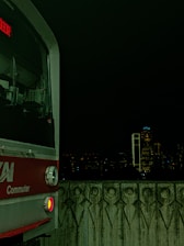 A commuter train is visible on the left side against a dark nighttime cityscape. The train has a red and silver exterior with a digital display showing text in red. The foreground features a patterned concrete barrier, and in the background, city buildings are faintly illuminated with a few bright lights.