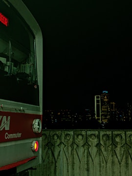 A commuter train is visible on the left side against a dark nighttime cityscape. The train has a red and silver exterior with a digital display showing text in red. The foreground features a patterned concrete barrier, and in the background, city buildings are faintly illuminated with a few bright lights.
