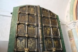 An ornate, ancient door featuring intricately carved religious icons in a grid pattern. The door frame is green with dark panels, each containing gold-painted depictions of various religious figures and scenes. The background includes a light-colored wall and an arch with decorative patterns.
