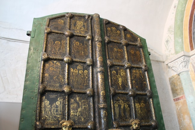 An ornate, ancient door featuring intricately carved religious icons in a grid pattern. The door frame is green with dark panels, each containing gold-painted depictions of various religious figures and scenes. The background includes a light-colored wall and an arch with decorative patterns.