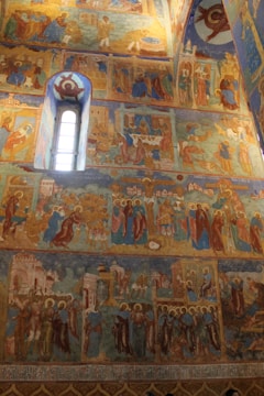 The image features a richly decorated wall inside an Orthodox church or monastery, covered with detailed frescoes. The frescoes depict biblical and religious scenes with numerous human figures, saints with halos, and architectural elements. The style is traditional, with vibrant colors and intricate patterns.