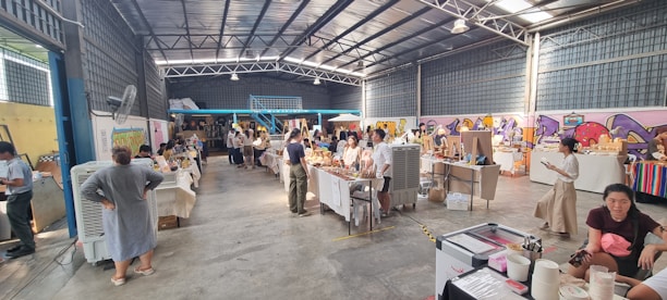 A bustling indoor market with several stalls and tables displaying various goods. People are browsing and interacting with vendors. The space is large and industrial with a high ceiling and visible beams. Walls are decorated with colorful graffiti art.
