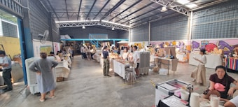 A bustling indoor market with several stalls and tables displaying various goods. People are browsing and interacting with vendors. The space is large and industrial with a high ceiling and visible beams. Walls are decorated with colorful graffiti art.