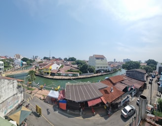 A vibrant urban landscape featuring a river canal surrounded by buildings with red-tiled roofs. The scene includes a few colorful tents lining the riverbank, lush green trees, and a bright blue sky with scattered clouds. Vehicles and pedestrians can be seen on the road adjacent to the river.