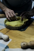 Hands are shaping a yellow dough inside a round, fluted tart pan on a wooden surface. Rolling pins and pastry tools are visible on the table, suggesting a baking activity.