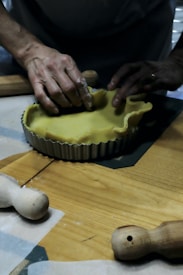 Hands are shaping a yellow dough inside a round, fluted tart pan on a wooden surface. Rolling pins and pastry tools are visible on the table, suggesting a baking activity.
