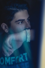 A person is gazing thoughtfully to the side, partially covering their face with an arm, creating a sense of introspection. The lighting is low, with a soft blue hue dominating the scene.