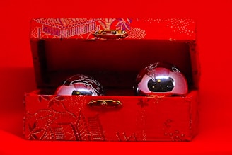 A decorative red box with intricate patterns is open, displaying two metallic balls inside. The exterior and interior textures of the box feature detailed designs, while the shiny balls reflect the surrounding light.