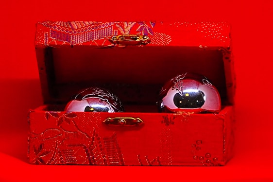 A decorative red box with intricate patterns is open, displaying two metallic balls inside. The exterior and interior textures of the box feature detailed designs, while the shiny balls reflect the surrounding light.