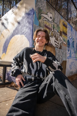 A person is sitting on the ground, laughing joyfully. The individual is wearing a patterned sweater and dark jeans. Behind them is a colorful mural on a wall, featuring an abstract design with a skeletal animal figure. Sunlight is casting shadows on the wall, creating an interplay of light and texture.