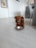 A small brown dog with long fur sits on a light wooden floor next to a metal bowl partially filled with dog food. The setting appears to be a cozy home interior with white walls, a gray couch, and a table visible in the background.