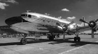 A vintage propeller airplane with a polished metal exterior is parked on an airfield. The aircraft has visible branded text on its side and is situated under a partly cloudy sky. The scene hints at an aviation event with fencing and a crowd visible in the background.
