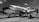 A vintage propeller airplane with a polished metal exterior is parked on an airfield. The aircraft has visible branded text on its side and is situated under a partly cloudy sky. The scene hints at an aviation event with fencing and a crowd visible in the background.