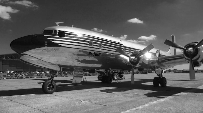 A vintage propeller airplane with a polished metal exterior is parked on an airfield. The aircraft has visible branded text on its side and is situated under a partly cloudy sky. The scene hints at an aviation event with fencing and a crowd visible in the background.