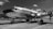 A vintage propeller airplane with a polished metal exterior is parked on an airfield. The aircraft has visible branded text on its side and is situated under a partly cloudy sky. The scene hints at an aviation event with fencing and a crowd visible in the background.