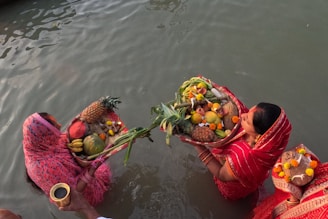Two women dressed in traditional clothing are partially submerged in water, holding baskets filled with fruits, coconuts, and marigold flowers. They appear to be participating in a ceremonial or religious ritual.