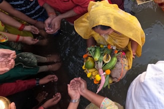 A group of people gathered around a body of water engage in a traditional ritual. A woman dressed in a yellow sari holds a decorated basket containing various fruits and vegetables, including bananas and coconuts, adorned with flowers. Hands of several participants are visible, cupped and extending towards the central figure in a gesture of worship or offering.