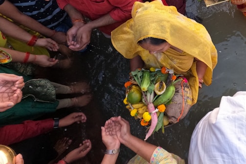A group of people gathered around a body of water engage in a traditional ritual. A woman dressed in a yellow sari holds a decorated basket containing various fruits and vegetables, including bananas and coconuts, adorned with flowers. Hands of several participants are visible, cupped and extending towards the central figure in a gesture of worship or offering.