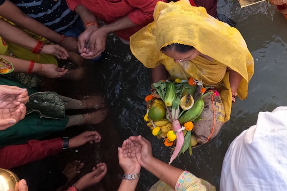 A group of people gathered around a body of water engage in a traditional ritual. A woman dressed in a yellow sari holds a decorated basket containing various fruits and vegetables, including bananas and coconuts, adorned with flowers. Hands of several participants are visible, cupped and extending towards the central figure in a gesture of worship or offering.