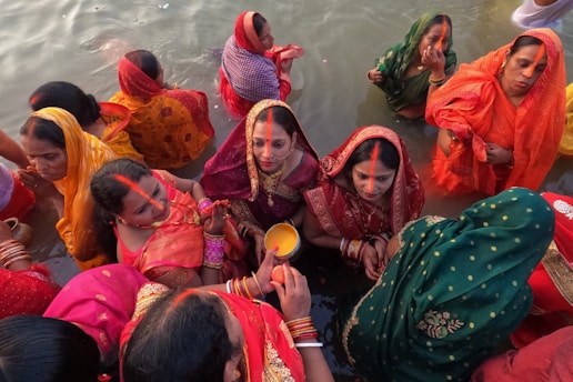 Women dressed in colorful traditional sarees are gathered in a body of water, likely participating in a religious or cultural ceremony. The women wear red, green, orange, and yellow garments with matching head coverings and jewelry. Several are holding offerings, while others have their hands in prayer or touching their foreheads.