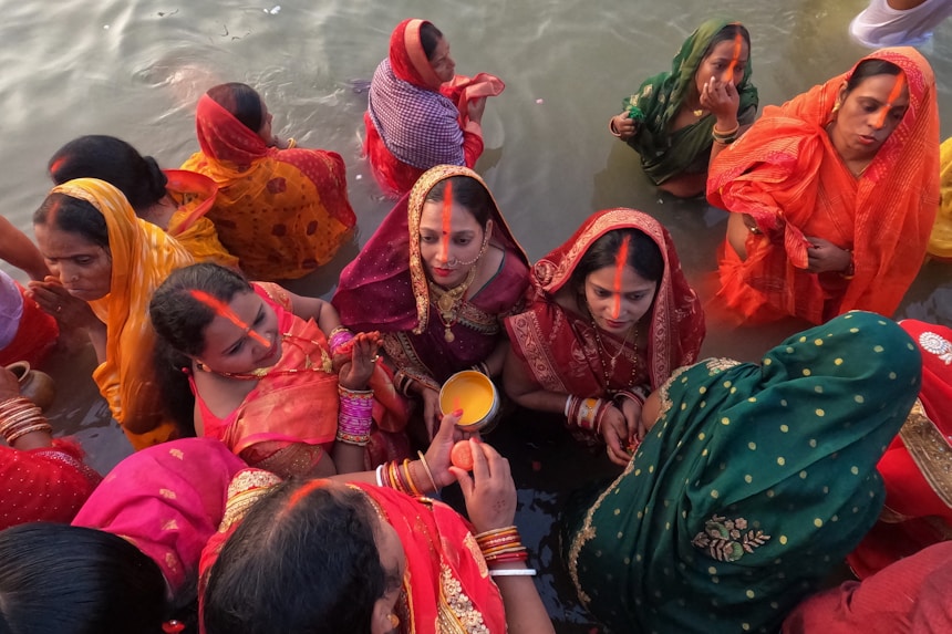 Women dressed in colorful traditional sarees are gathered in a body of water, likely participating in a religious or cultural ceremony. The women wear red, green, orange, and yellow garments with matching head coverings and jewelry. Several are holding offerings, while others have their hands in prayer or touching their foreheads.